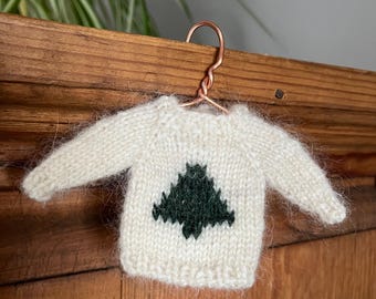 May include: A miniature cream-colored knitted sweater featuring a dark green pine tree graphic on the front. The sweater is suspended from a small, twisted copper-colored wire hanger, showcasing a fuzzy texture and ribbed cuffs and neckline.