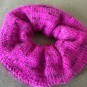 May include: A bright fuchsia pink hair scrunchie made of a knitted fabric with a fuzzy texture. The circular accessory features a gathered, pleated design with subtle dark speckles woven throughout the vibrant pink yarn, creating a soft appearance.
