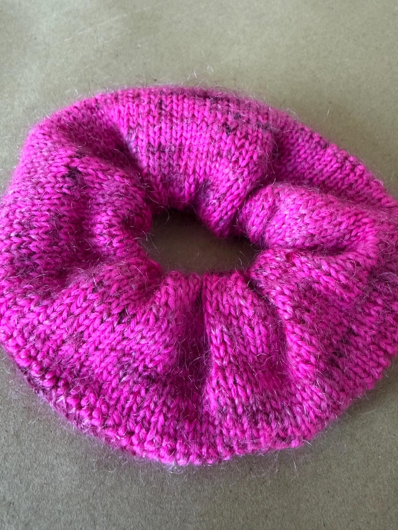 May include: A bright fuchsia pink hair scrunchie made of a knitted fabric with a fuzzy texture. The circular accessory features a gathered, pleated design with subtle dark speckles woven throughout the vibrant pink yarn, creating a soft appearance.