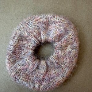 May include: A circular hair scrunchie made of a fuzzy, knitted yarn featuring a variegated color palette of soft pink, lavender, and cream tones. The accessory has a gathered, elasticized construction with a visible, textured, and soft fiber finish.
