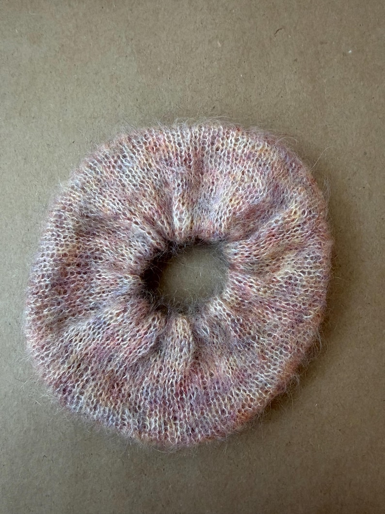 May include: A circular hair scrunchie made of a fuzzy, knitted yarn featuring a variegated color palette of soft pink, lavender, and cream tones. The accessory has a gathered, elasticized construction with a visible, textured, and soft fiber finish.
