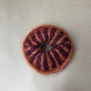 May include: A circular hair scrunchie featuring a fuzzy, knitted texture with alternating vertical stripes in deep purple and burnt orange. The accessory has a gathered, donut-like shape with a central opening, showcasing a soft, fibrous yarn construction.