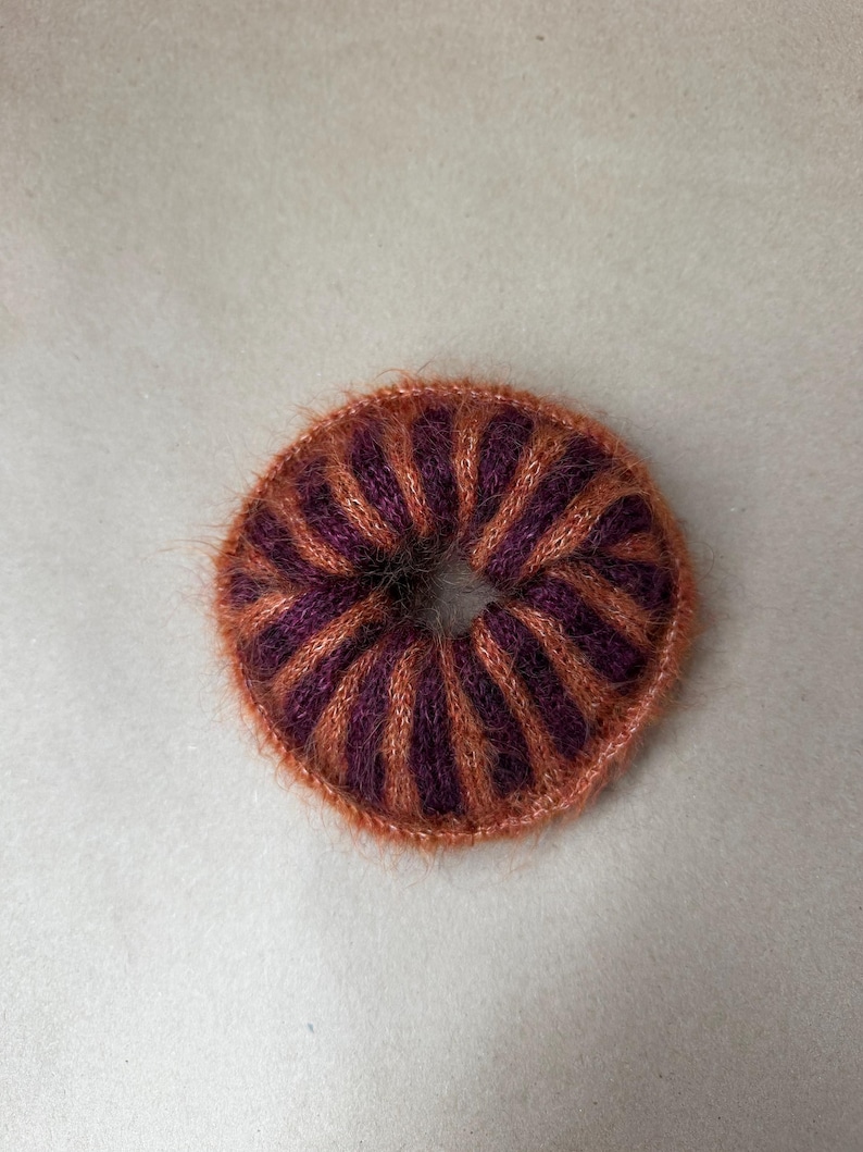 May include: A circular hair scrunchie featuring a fuzzy, knitted texture with alternating vertical stripes in deep purple and burnt orange. The accessory has a gathered, donut-like shape with a central opening, showcasing a soft, fibrous yarn construction.