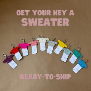 May include: A collection of keychains shaped like miniature sweaters in various colors, including red, pink, and blue. Each keychain has a metal clasp and a small tag. The text "GET YOUR KEY A SWEATER" and "READY-TO-SHIP" are displayed.