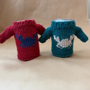 May include: Two knitted can koozies, one red with a blue design and the other teal with a white design. Each koozie is shaped like a tiny sweater with sleeves. The koozies are designed to keep beverages cold and add a touch of whimsy.