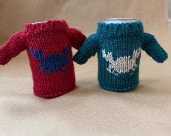 May include: Two knitted can koozies, one red with a blue design and the other teal with a white design. Each koozie is shaped like a tiny sweater with sleeves. The koozies are designed to keep beverages cold and add a touch of whimsy.