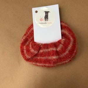 May include: A fuzzy, red and white striped knit scrunchie attached to a white rectangular display card. The card features a circular logo with a grey dog illustration, a pink ball of yarn, and the text farndoodle knits printed in black and red.