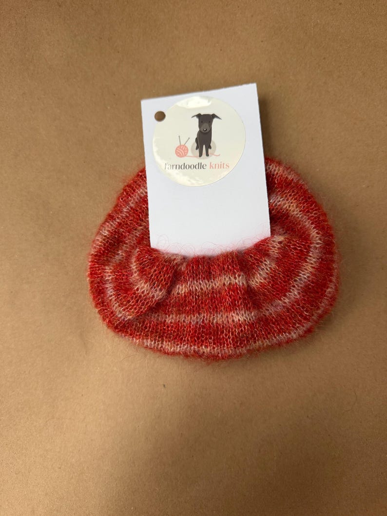 May include: A fuzzy, red and white striped knit scrunchie attached to a white rectangular display card. The card features a circular logo with a grey dog illustration, a pink ball of yarn, and the text farndoodle knits printed in black and red.