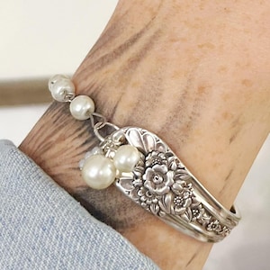 May include: A silver spoon bracelet with floral detailing and dangling faux pearls. The bracelet is worn on a wrist, showcasing the intricate design and vintage aesthetic. The spoon's handle is bent to form the bracelet.