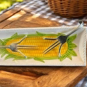 May include: A rectangular white ceramic serving dish with a corn on the cob design and two metal forks. The dish is on a wooden cutting board, with two wicker baskets filled with fruit in the background. The table is covered with a checkered tablecloth.