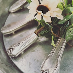 May include: Silverware with ornate designs, arranged in a metal bowl with a white flower and green leaves.