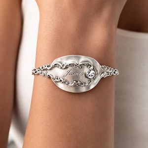 May include: A silver spoon bracelet with the name "James" engraved on the spoon's bowl. A clear gemstone is set near the name. The bracelet has ornate detailing and is worn on a wrist.