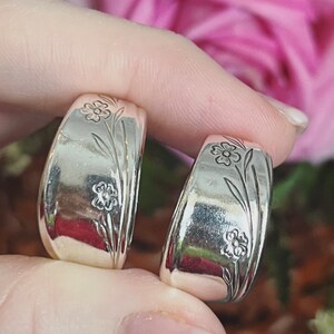 May include: Two silver hoop earrings with a floral design. The earrings are made from recycled silverware and feature a delicate floral pattern.