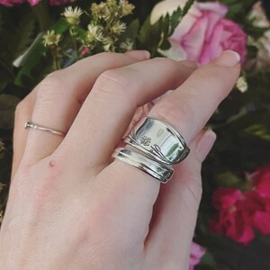 K&ouml;nnte beinhalten: Ein silberner L&ouml;ffelring mit floralem Design. Der Ring wird an einem Finger mit einem schlichten Silberband getragen.