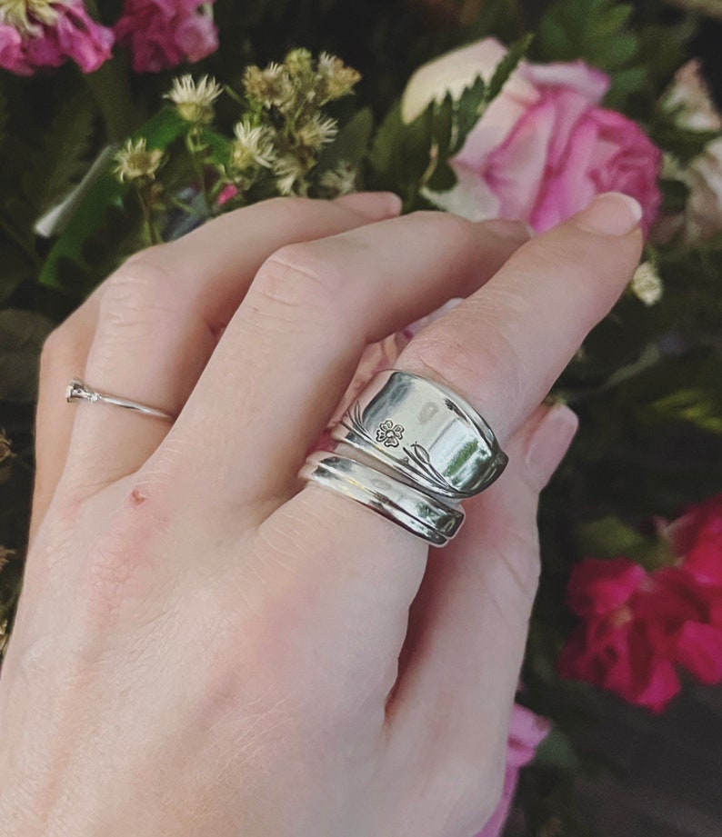K&ouml;nnte beinhalten: Ein silberner L&ouml;ffelring mit floralem Design. Der Ring wird an einem Finger mit einem schlichten Silberband getragen.