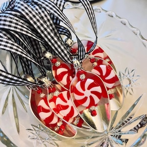 May include: A set of silver spoons with red and white peppermint candy ornaments attached. The spoons are tied together with black and white checkered ribbon.