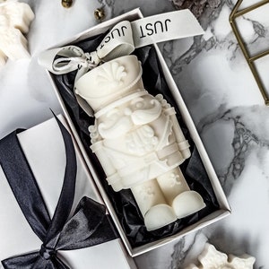 Può includere: Una candela in ceramica bianca a forma di schiaccianoci in una scatola regalo bianca con un nastro nero. La candela è a forma di schiaccianoci tradizionale con un cappello rosso e una barba bianca. La scatola è legata con un nastro nero e ha la scritta "Just" stampata.
