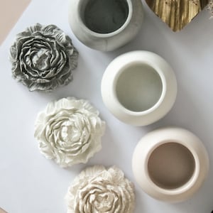 May include: An overhead shot of several decorative items. There are three small, round, open-topped vases in shades of grey and cream. Three flower-shaped wall decorations are also present, in matching colours. The items are arranged on a white surface.