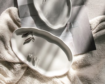 May include: Two white ceramic trays with irregular edges. The larger tray has a gold beaded bracelet and two silver hair clips. The smaller tray has two silver earrings and a clear crystal ring.