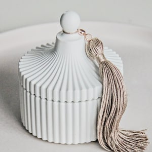 May include: White ceramic candle jar with a ribbed design and a dome-shaped lid. The jar has a tassel attached to the lid.
