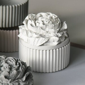 May include: Two white ceramic containers with a decorative flower design on the lid. The containers have a ribbed texture and are perfect for storing small items.