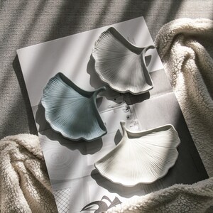 May include: Three ceramic dishes shaped like ginkgo leaves. The dishes are in shades of white, light blue, and grey. The dishes are arranged on a white surface with a patterned background.