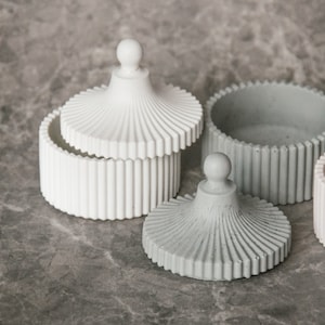 May include: Two decorative containers with lids. The larger container is white with a ribbed design and the smaller container is gray with a ribbed design.