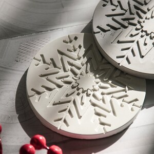 May include: Two white ceramic coasters with a snowflake design. The coasters are round and have a raised snowflake pattern.