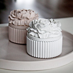 Puede incluir: Dos cajas decorativas con diseños florales. La caja de la izquierda es de color rosa claro con un diseño de flor rosa. La caja de la derecha es blanca con un diseño de flor blanca. Ambas cajas tienen una textura acanalada.