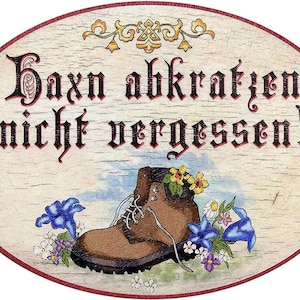 May include: Oval wooden sign with the German text "Boxn abkratzen nicht vergessen!" above a brown hiking boot filled with yellow flowers. Blue and white flowers surround the boot, with a red border and decorative flourishes.