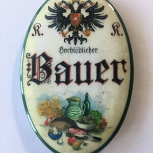 Op de afbeelding: Een ovaalvormig bord met een groene rand en een witte achtergrond. Het bord toont een zwarte dubbelkoppige adelaar met een rood schild en gouden accenten. De tekst "Hochlöblicher Bauer" is geschreven in zwarte cursieve letters. De onderkant van het bord toont een stilleven met brood, groenten, eieren en een mes.