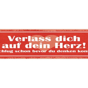 Może przedstawiać: Czerwony prostokątny znak z białym tekstem, który brzmi "Verlass dich auf dein Herz!" i "Es schlug schon bevor du denken konntest." Znak ma zużyty wygląd i grafikę różowego serca.