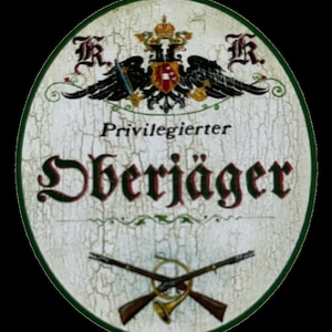 Puede incluir: Un letrero ovalado blanco con borde verde y acabado desgastado. El letrero presenta un águila negra con una corona y un escudo, junto con el texto "Privilegierter" y "Dherjäger" en una fuente decorativa.