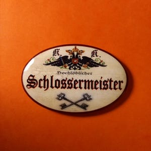 Puede incluir: Letrero ovalado con la palabra alemana "Schlossermeister" en una fuente decorativa. El letrero presenta un escudo con un águila bicéfala y llaves cruzadas. El fondo es blanquecino con un aspecto envejecido, sobre un fondo naranja.