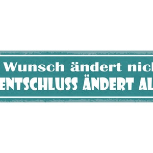 May include: A teal blue rectangular sign with white text that reads "Ein Wunsch ändert nichts. EIN ENTSCHEIDUNG ÄNDERT ALLES!"