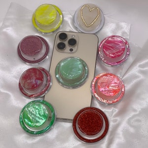 May include: Assortment of phone grips in various colors and patterns, including glitter, pearlescent, and solid colors. The grips are designed to be attached to the back of a phone for a secure hold.