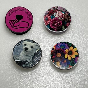 May include: Four different phone pop sockets. One is pink with a hand holding a heart and the text "#LittleThings HandmadeByBabs". One is white with a floral design featuring pink, red, and white roses. One is black with a photo of a white dog. One is white with a floral design featuring yellow, purple, and white flowers.