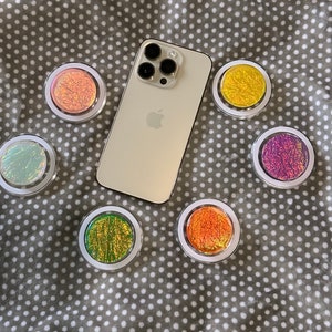 May include: A white iPhone with a black camera lens and a white background. The phone is surrounded by six colorful phone grips with iridescent finishes.