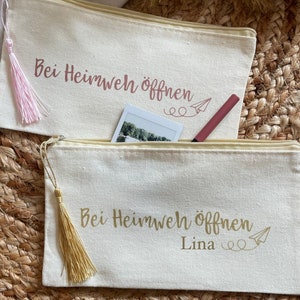 Könnte beinhalten: Zwei weiße Canvas-Reißverschlusstaschen mit goldenen und rosafarbenen Quasten. Die Taschen haben den Text "Bei Heimweh öffnen" in goldener und rosafarbener Schrift, mit einer Papierflugzeuggrafik. Die untere Tasche hat den Namen "Lina" unter dem Text geschrieben.