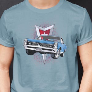 May include: A blue t-shirt with a vintage Pontiac GTO muscle car graphic. The car is blue with a red and white Pontiac logo. The shirt is a light blue color.