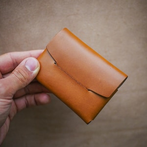 May include: A small, tan leather wallet with a flap closure. The wallet is rectangular and appears to be handmade, with visible stitching and a simple design. The leather has a smooth texture and a warm, natural color.