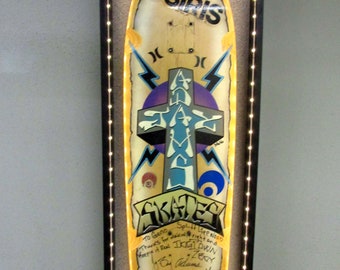 Könnte beinhalten: Ein gelbes und blaues Skateboard-Deck mit einem Kreuzdesign und dem aufgedruckten Wort "OSIRIS". Das Deck ist in einem schwarzen Rahmen mit einem weißen Lichtrahmen gerahmt. Das Deck ist von mehreren Personen signiert, darunter "Dogtown" und "Zegy Adams".
