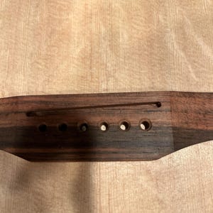 May include: Close-up of a wooden acoustic guitar. The bridge is dark brown with six holes for strings. The soundhole is framed with a decorative inlay. The wood grain of the guitar body is visible.