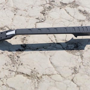 May include: A grey guitar neck with a white headstock overlay. The neck features frets and tuning peg holes. The guitar neck is set against a textured, light-coloured background.