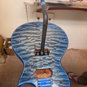 May include: An unfinished electric guitar body featuring a blue quilted maple top with a wavy pattern. The body has two rectangular pickup cavities, several circular holes for hardware, and a blue tape covering the neck joint area.