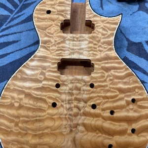 May include: Unfinished electric guitar body with a figured maple top. The wood grain has a wavy pattern in shades of light and medium brown. The body has cutouts for the neck, pickups, and controls, and several drilled holes.