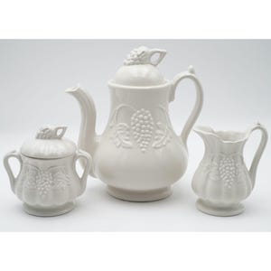 May include: A white ceramic tea set comprising a teapot, sugar bowl, and a creamer. Each item is decorated with a grape cluster motif. The teapot has a lid with a grape cluster handle, and the creamer has a curved handle.
