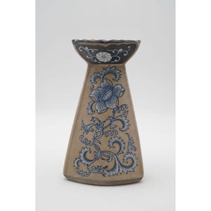 May include: A beige ceramic vase with a flared, trapezoidal shape. The vase has a dark blue rim and is decorated with blue floral and vine patterns. The design includes a large flower and scrollwork, creating an elegant aesthetic.