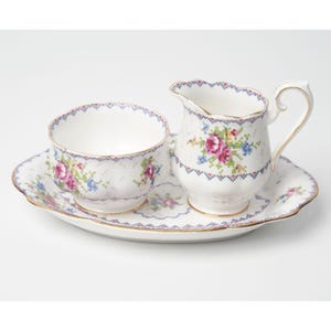 May include: A vintage porcelain tea set featuring a cup, creamer, and serving tray. The white set is decorated with a floral cross-stitch pattern in pink, blue, and green, with gold trim along the edges. The tray is oval-shaped.