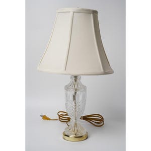 Może przedstawiać: Lampa stołowa z przezroczystą, szlifowaną szklaną podstawą w romby oraz okrągłą podstawką z metalu w kolorze złota. Lampa posiada zwężający się abażur z tkaniny w kolorze złamanej bieli oraz brązowy przewód elektryczny z żółtą wtyczką.